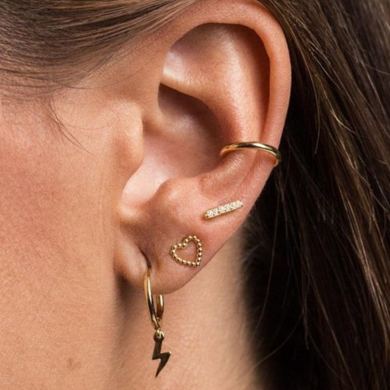 A woman wearing gold heart studs, ear cuff, and lightning bolt huggies.