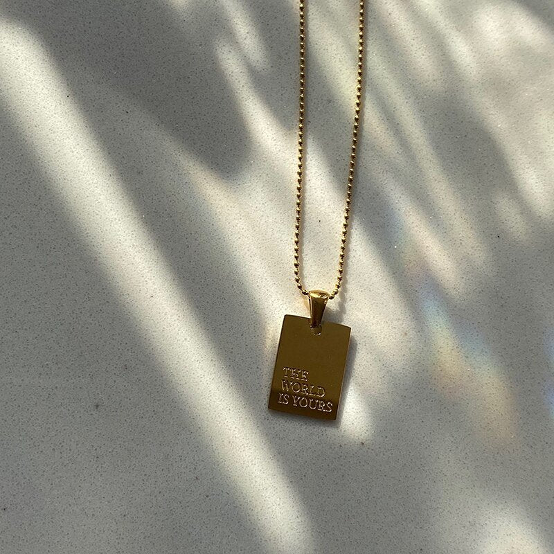 Gold dog tag necklace with "The World Is Yours" engraved on it.