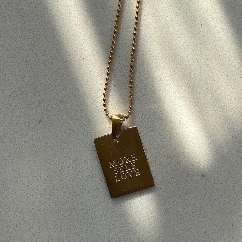 Gold dog tag necklace with "More Self Love" engraved on it.